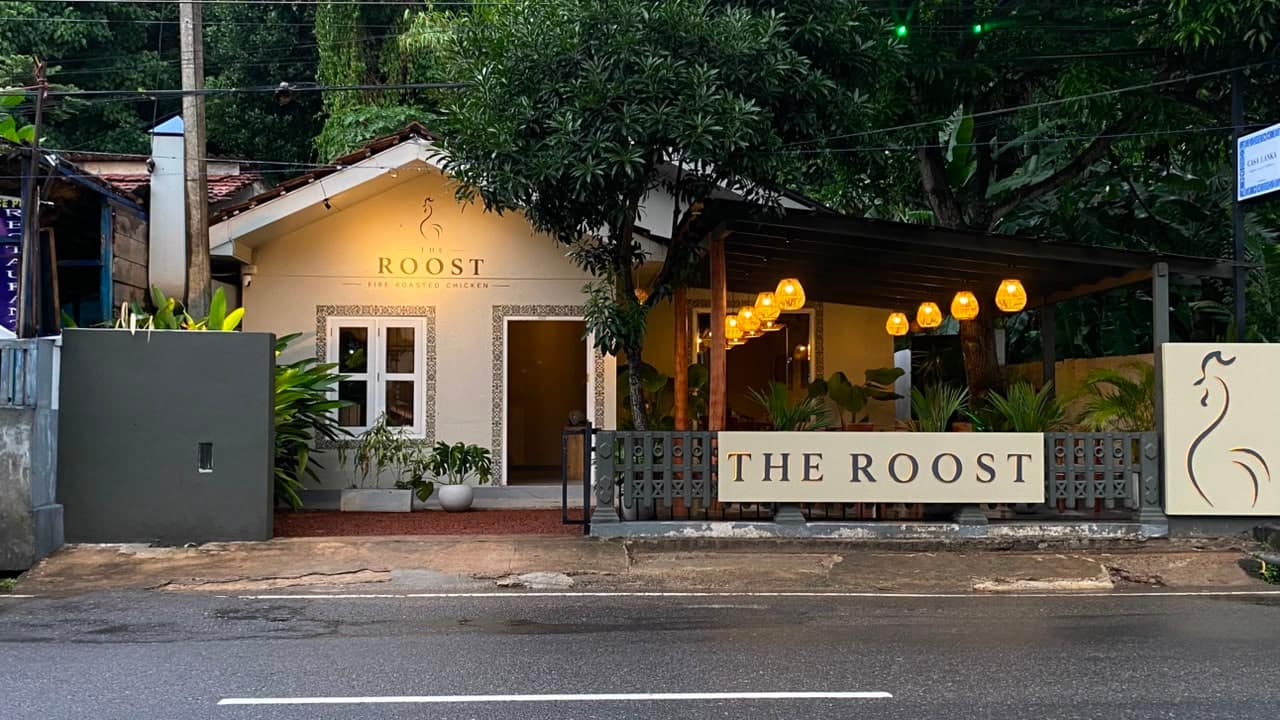 The Roost from the road at dusk — cream sign on the grey fence, lanterns lit under the canopy