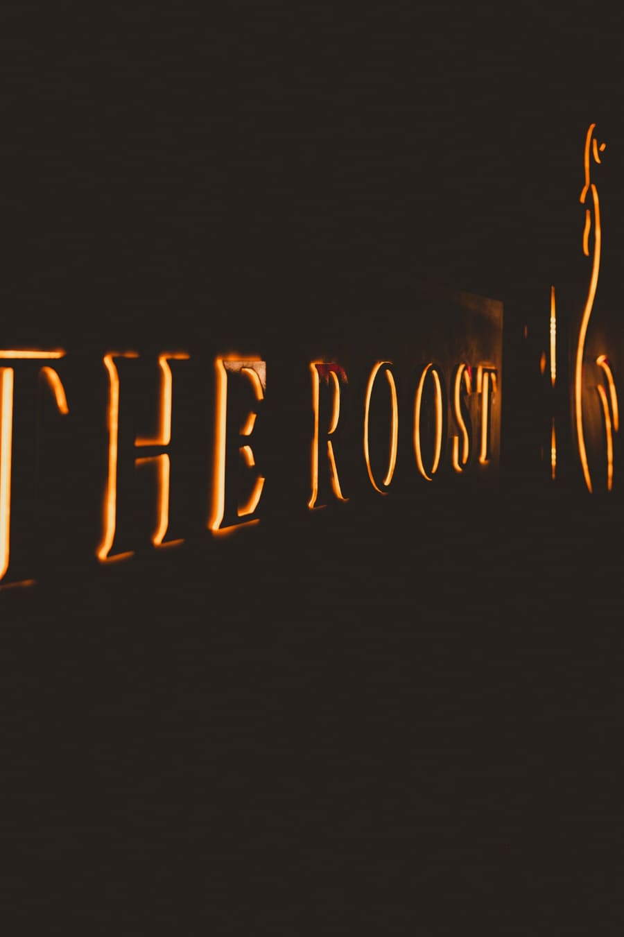The Roost sign at night, letters and rooster backlit in fire orange